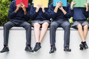 four students in school uniform sitting on a wall holding up different coloured books in front of their faces
