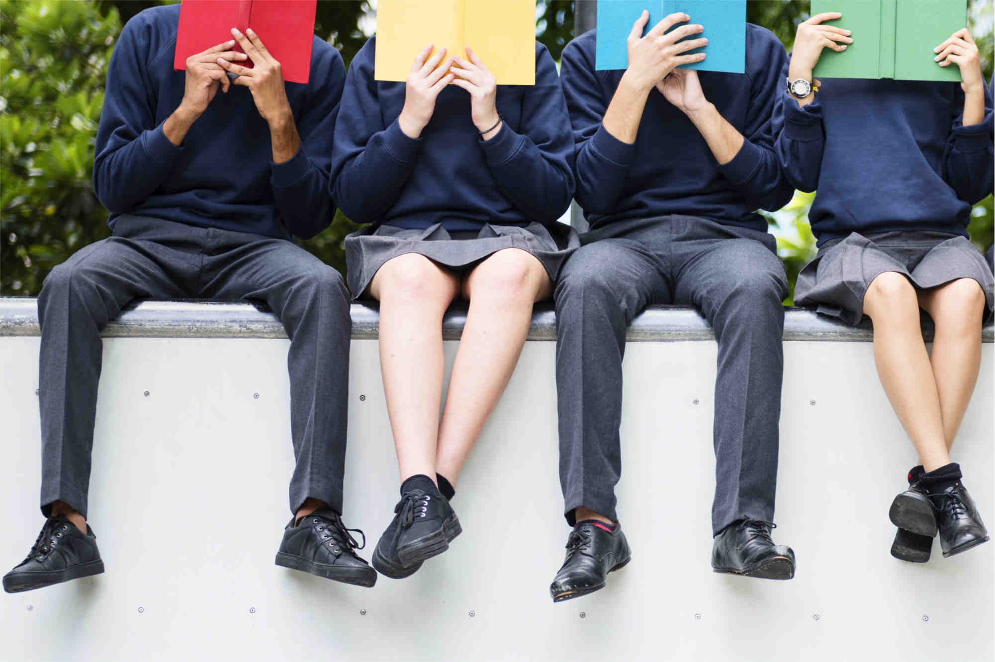four students in school uniform sitting on a wall holding up different coloured books in front of their faces  - Footsteps Educational Psychology Assessment and Advice