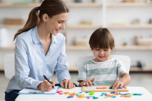 woman in blue shirt and child in green and white striped tee-shirt sitting at classroom desk child is placing magnetic letters on small whiteboard on desk, woman is writing, both smiling and looking at whiteboard