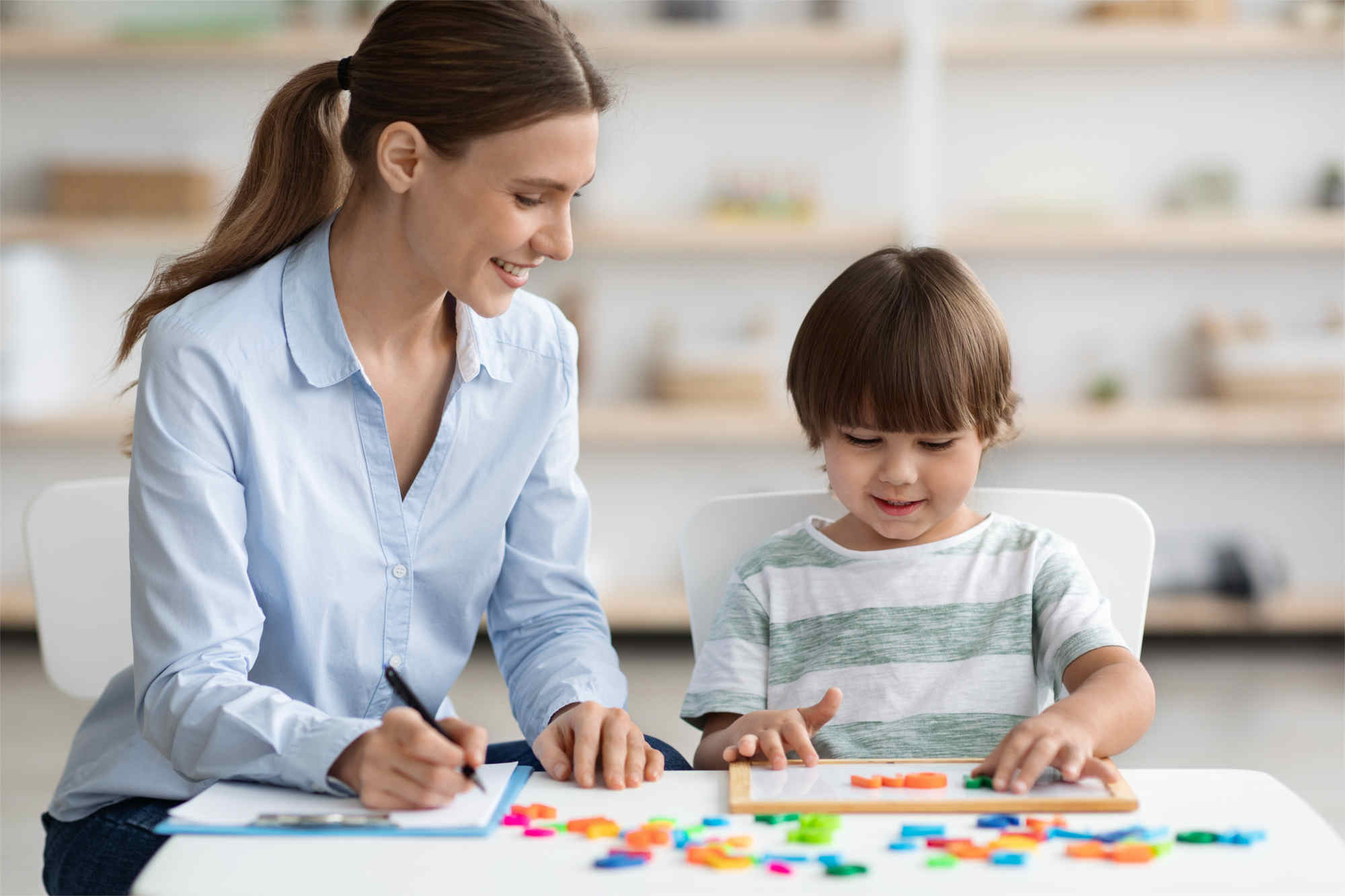 woman in blue shirt and child in green and white striped tee-shirt sitting at classroom desk  child is placing magnetic letters on small whiteboard on desk, woman is writing, both smiling and looking at whiteboard - Footsteps Educational Psychology Assessment and Intervention