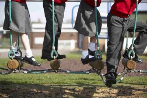 bottom half of four children wearing uniform of red tops and grey skirts or trousers walking on log and rope bridge in playground school out of focus in background
