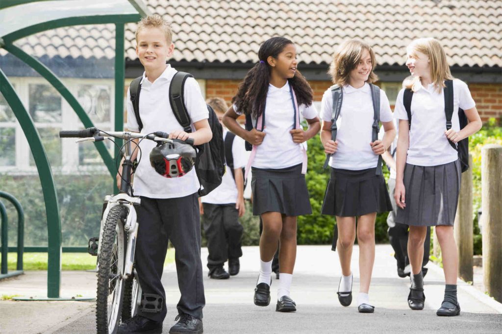 children in white polo shirts and grey trousers or skirts school uniform near school bike shelter a boy in the the foreground with his bike and three girls walking and smiling as they talk to each other